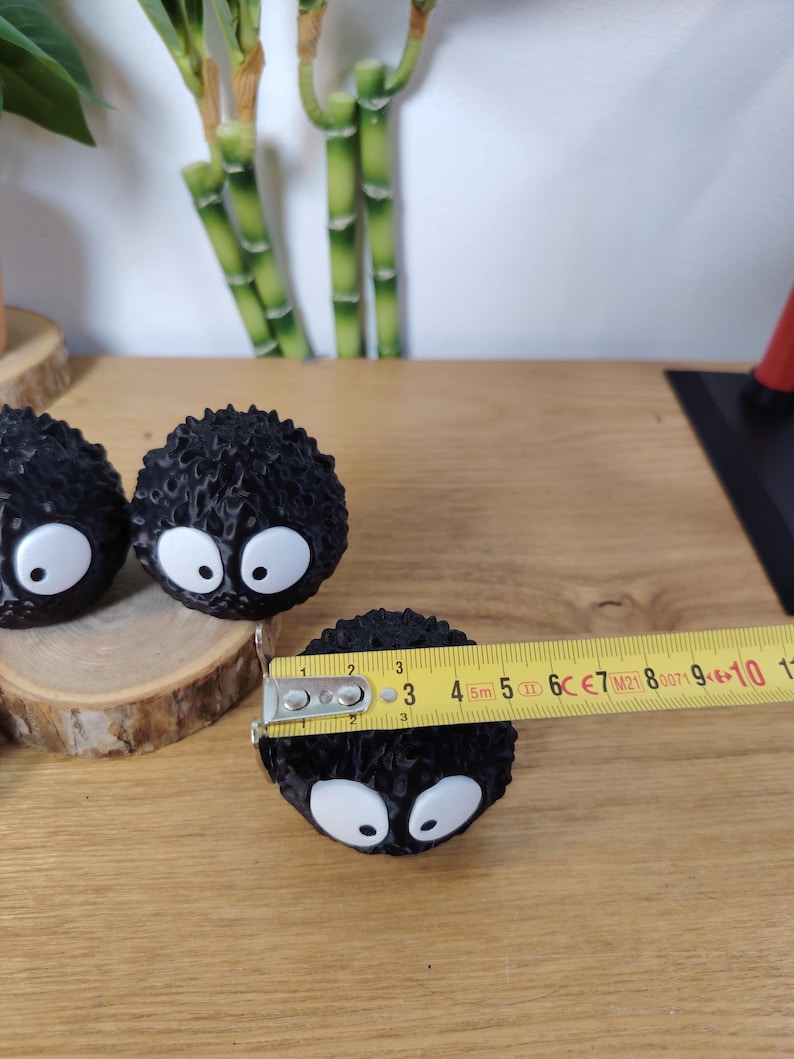 May include: Three black, round, spiky figurines with large white eyes. A measuring tape shows one figurine is approximately 5 cm in diameter. The figurines are on a wooden surface.