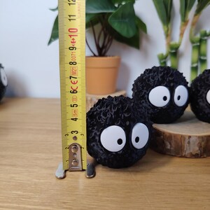 May include: Black, round, fuzzy decorative figures with large white eyes, resembling soot sprites. The figures are approximately 7.6 cm in height. They are displayed on a wooden surface with a measuring tape in the background.