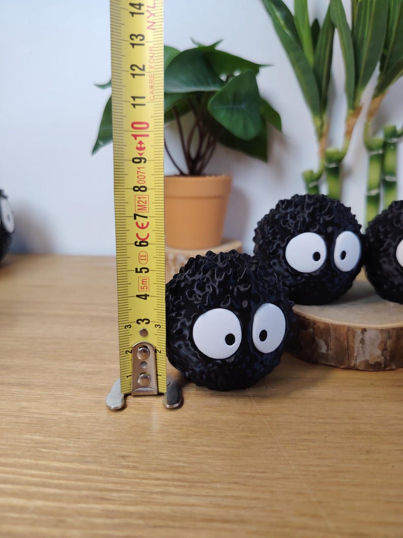 May include: Black, round, fuzzy decorative figures with large white eyes, resembling soot sprites. The figures are approximately 7.6 cm in height. They are displayed on a wooden surface with a measuring tape in the background.
