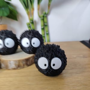 May include: Three black, round, fuzzy figurines with large white eyes and black pupils. The figurines are arranged on a wooden surface, with bamboo stalks in the background. The figurines resemble soot sprites.