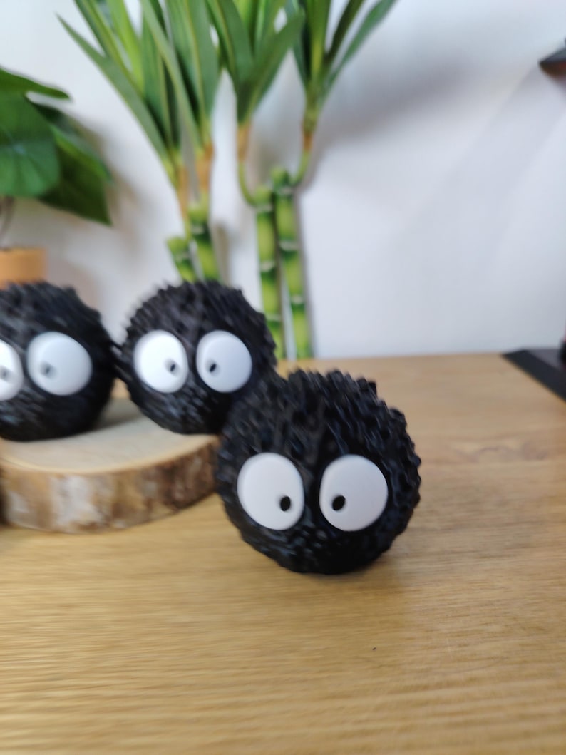 May include: Three black, round, fuzzy figurines with large white eyes and black pupils. The figurines are arranged on a wooden surface, with bamboo stalks in the background. The figurines resemble soot sprites.