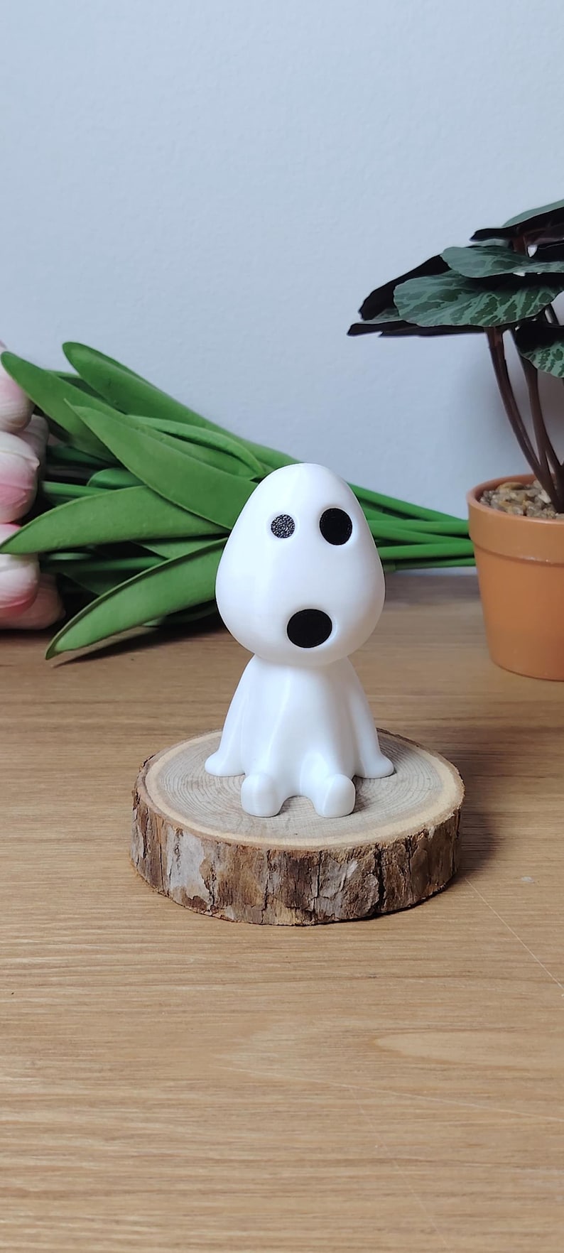 K&ouml;nnte beinhalten: Wei&szlig;e 3D-gedruckte Figur eines Charakters mit einem runden Kopf, zwei schwarzen Augen und einem schwarzen Mund. Die Figur sitzt auf einer Holzscheibe.