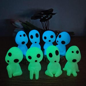 May include: A group of eight glow-in-the-dark figurines. The figurines are shaped like small, round people with large eyes and simple features. They are in two colors: blue and green.