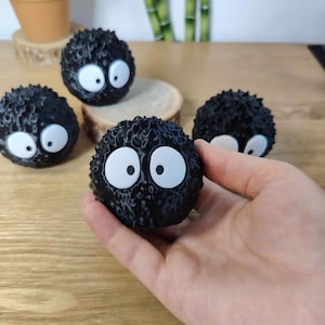 May include: A hand holds a black, round, fuzzy character with large white eyes and black pupils. Several similar characters are scattered around on a wooden surface. The characters have a spiky texture and a cartoonish appearance.