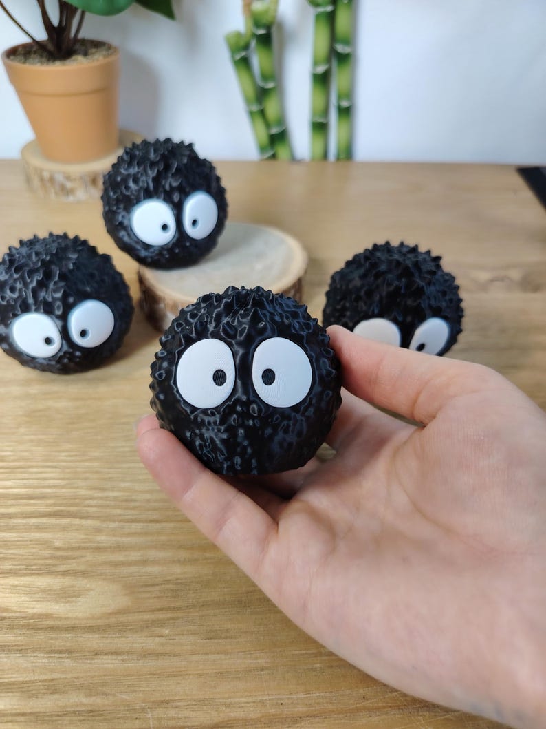 May include: A hand holds a black, round, fuzzy character with large white eyes and black pupils. Several similar characters are scattered around on a wooden surface. The characters have a spiky texture and a cartoonish appearance.