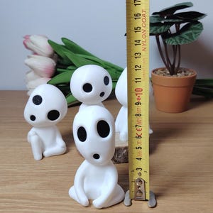 K&ouml;nnte beinhalten: Vier wei&szlig;e Figuren mit schwarzen Augen und M&uuml;ndern, die an Figuren aus dem Animationsfilm "Mein Nachbar Totoro" erinnern. Die Figuren sitzen auf einer Holzoberfl&auml;che, im Hintergrund befindet sich ein Ma&szlig;band.