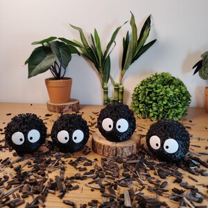 May include: Four black, round, fuzzy figures with large white eyes, resembling soot sprites. They are displayed on a wooden surface scattered with wood chips. Green plants in pots and a decorative bush are in the background. The figures are a popular animated film character.