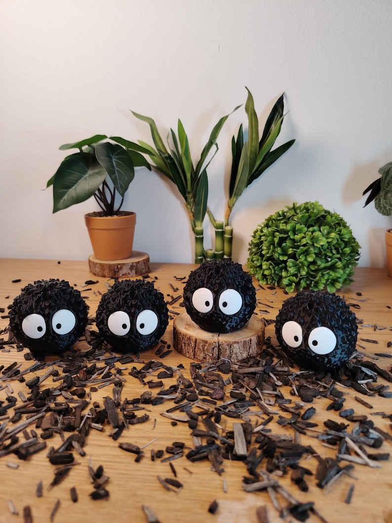 May include: Four black, round, fuzzy figures with large white eyes, resembling soot sprites. They are displayed on a wooden surface scattered with wood chips. Green plants in pots and a decorative bush are in the background. The figures are a popular animated film character.