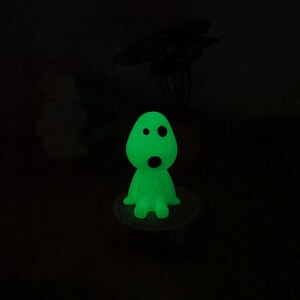 May include: A green glow-in-the-dark figurine of a ghost with two eyes and a mouth. The ghost is sitting on a small white base.