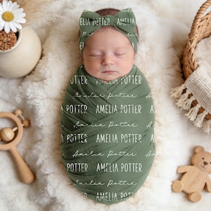 May include: A newborn baby wrapped in a green swaddle blanket and matching headband, both printed with the name "Amelia Potter" in white script. The baby is lying on a fluffy white surface, with a wooden rattle and a small wooden bear toy nearby.