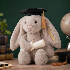 Puede incluir: Un conejito de peluche gris claro con un birrete negro y una borla dorada. El conejito sostiene un pergamino enrollado y tiene el nombre "Sophia" bordado en una oreja. El conejito está sentado sobre una superficie de madera.