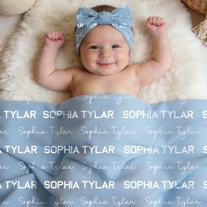 Op de afbeelding: Een baby met een blauwe haarband ligt op een witte, zachte vloer. De baby is bedekt met een lichtblauwe deken bedrukt met de naam "Sophia Tylar" in het wit. De haarband heeft ook de naam erop.