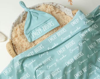 May include: A light blue baby blanket and matching knotted hat, both with the name "EMERY BROOKS" printed in white. The items are displayed in a woven basket with a soft, beige lining.