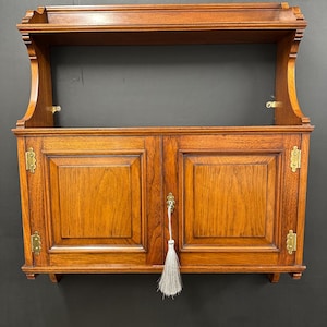 Puede incluir: Un mueble de madera para colgar en la pared con dos puertas y una balda. El mueble es de madera de tonos cálidos, con herrajes de latón y una borla decorativa. El diseño es clásico y elegante, ideal para almacenamiento.