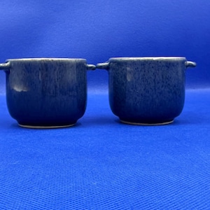 May include: Two matching blue ceramic bowls with small handles. The bowls have a speckled texture and a glossy finish. The bowls are sitting on a blue surface. The bowls are likely for serving soup or other dishes.