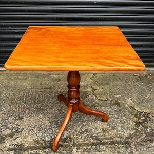 May include: A small, square, wooden table with a turned pedestal base and three legs. The table top is a rich, reddish-brown color.