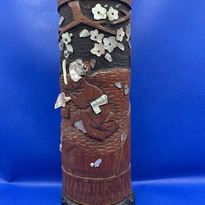 May include: A cylindrical brown wooden container featuring intricate carvings of a figure in traditional clothing and floral branches. The design is accented with inlaid iridescent mother-of-pearl pieces, and the base has a dark, textured carved border.