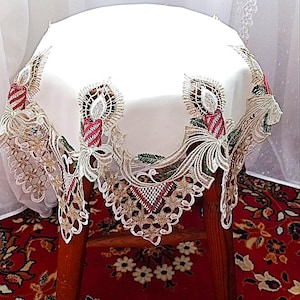Pode incluir: Toalha de mesa redonda branca com borda de renda intrincada com desenhos de velas em vermelho, dourado e verde. A toalha de mesa está sobre um banquinho de madeira, mostrando o bordado detalhado e as cores festivas.