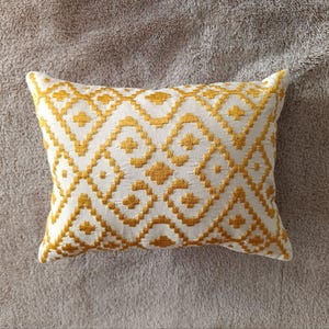 May include: Rectangular decorative pillow with a white background and a repeating geometric pattern in gold. The pattern consists of diamond and square shapes. The pillow is placed on a textured, neutral-colored surface.