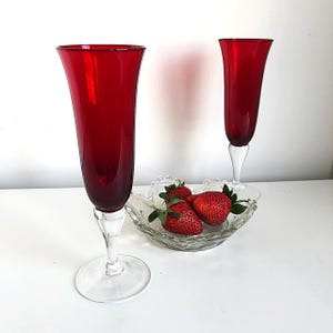 Peut inclure: Deux flûtes à champagne rouge rubis avec des pieds et des bases transparents se trouvent à côté d'un plat en verre contenant des fraises fraîches. Les flûtes ont un design classique et élégant, parfait pour les célébrations.