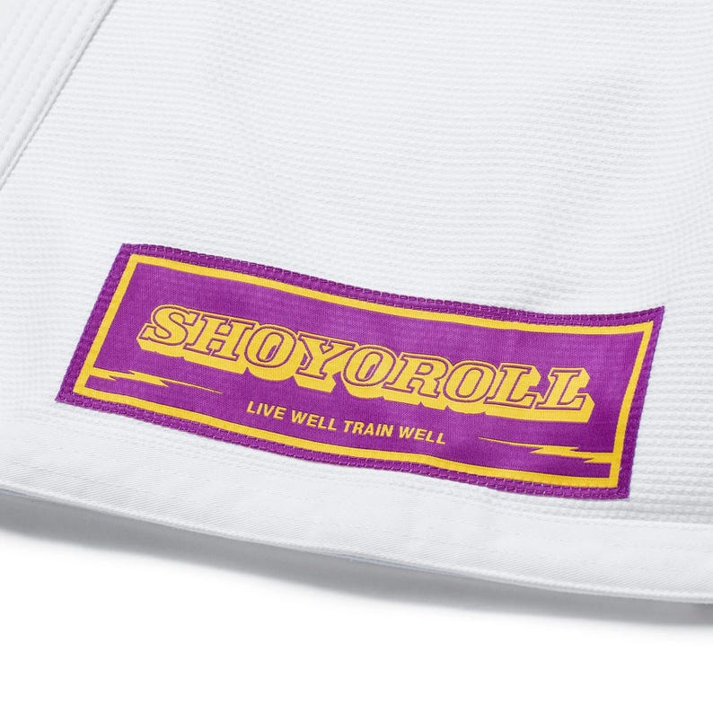 Puede incluir: Primer plano de un uniforme de artes marciales blanco con un parche rectangular morado y amarillo. El parche presenta la palabra "SHOYOROLL" en amarillo, con el texto "LIVE WELL TRAIN WELL" debajo.