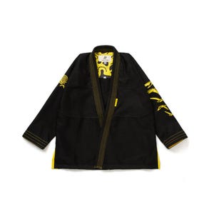 Op de afbeelding: Zwart martial arts uniform met gele accenten. De jas heeft gele afwerking langs de kraag, manchetten en zoom. Gele graphics staan op de mouwen en binnenkant van de kraag. Het uniform ligt plat op een witte achtergrond.