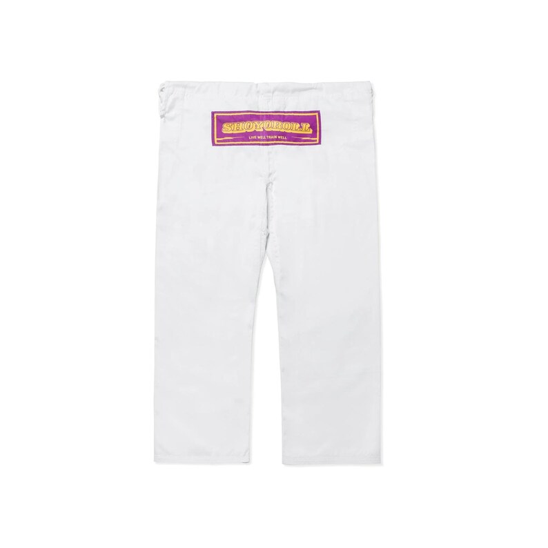 Puede incluir: Pantalones blancos con un parche rectangular morado y dorado. El parche tiene el texto "SHOW YOUR ROLL" y "LIVE WELL, PLAY WELL". Los pantalones tienen un dise&ntilde;o de pierna recta.