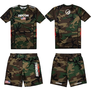 May include: A camouflage pattern athletic shirt and shorts set. The shirt has "DEFCON" and an American flag graphic. The shorts have "SHOYOROLL" printed vertically. Both pieces are in shades of green, brown, and black.
