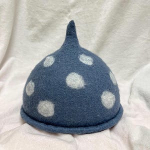 Può includere: Un cappello blu in feltro con pois bianchi e una parte superiore a punta. Il cappello ha una base arrotondata ed è posto su uno sfondo bianco strutturato. Il design del cappello è semplice e stravagante.