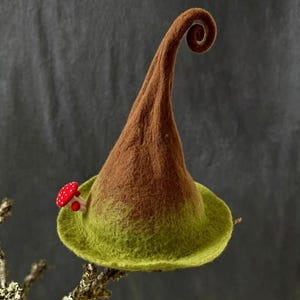 May include: A whimsical, handmade felt hat with a brown, curled tip and a green brim. A small, red mushroom with white spots accents the hat's side. The hat is designed to sit on the head and is perfect for costume or cosplay.