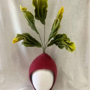 May include: A whimsical red bonnet with a radish design. The bonnet features a red cap with a green and yellow leafy top, and ties under the chin. The hat is displayed on a white mannequin head.