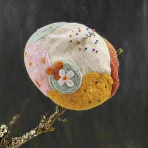 May include: A colorful, handmade felt beret. The beret features a patchwork design with sections of pink, orange, yellow, and green. It is adorned with small, colorful beads and a felt flower applique. The beret is displayed on a natural branch.