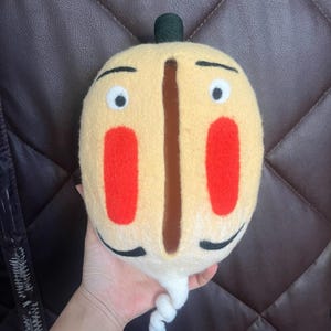 May include: A hand-felted plush toy shaped like a cartoon face with a long white tail. The face is yellow with red cheeks, black eyebrows, and large white eyes. The toy has a black top hat and a brown vertical line down the center of the face.