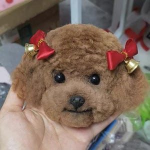 Può includere: Una testa di cane marrone e peluche con occhi neri e naso nero. Il cane ha due fiocchi rossi con campanelle dorate attaccate alle orecchie. Il peluche è progettato per assomigliare a un barboncino.