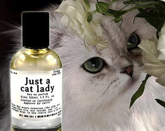 May include: A clear glass perfume bottle with a black cap and a white label reading Just a cat lady, Eau de parfum, Size: 50ml, 1.7 FL. oz, Crafted in California, approve by cat!!!, No. ∞, Friday the 13th, and additional descriptive text.