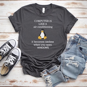 Può includere: T-shirt grigio scuro con la scritta "COMPUTER IS LIKE A air conditioning it becomes useless when you open WINDOWS" e una grafica a forma di pinguino. La maglietta è abbinata a jeans blu e scarpe da ginnastica nere.