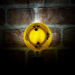 May include: A yellow night light with a decorative black floral design. The night light has a circular shape with two black dots and is set against a brick wall.