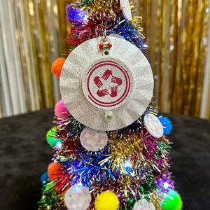May include: A white Christmas tree ornament with a red five-pointed star design. The star has five hands reaching out from the points. The ornament is hanging on a small, colorful Christmas tree.