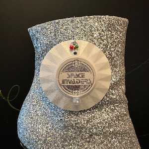 May include: A white, round, decorative ornament with a silver glitter finish. The ornament has a raised, circular design with the text "SPACE INVADERS" in black lettering. The ornament is hanging from a silver string with red and green beads.