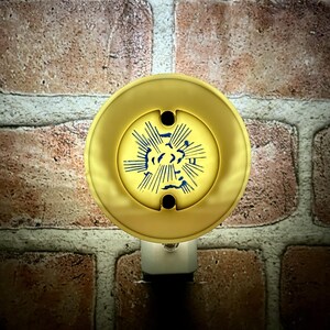 May include: A gold-colored night light with a circular design featuring blue lines radiating from a central shape. The night light is plugged into a white outlet and set against a brick wall.