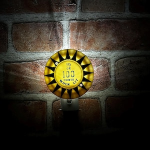 May include: A yellow and black night light with the text "10 OR 100 POINTS WHEN LIT". The night light has a circular design with a starburst pattern around the edge. It is plugged into a wall outlet against a brick wall.