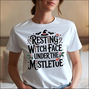 Könnte beinhalten: Weißes T-Shirt mit dem Text "Resting Witch Face Under The Mistletoe" in schwarzer Schrift. Das Design beinhaltet einen Hexenhut, Fledermäuse, Zuckerstangen und Stechpalmen, für eine festliche und humorvolle Atmosphäre.