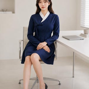 May include: A navy blue dress with long sleeves and a white V-neck collar. The dress features a wrap-style bodice with a tie closure at the waist, a fitted midsection, and a flared skirt that reaches above the knees. The fabric is solid navy blue.