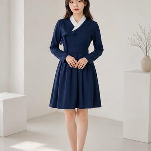 May include: A navy blue knee-length dress featuring a white V-neck collar, long sleeves, and a wrap-style bodice with a side tie closure. The skirt is pleated and flares from the waist, paired with black closed-toe pumps for a formal appearance.