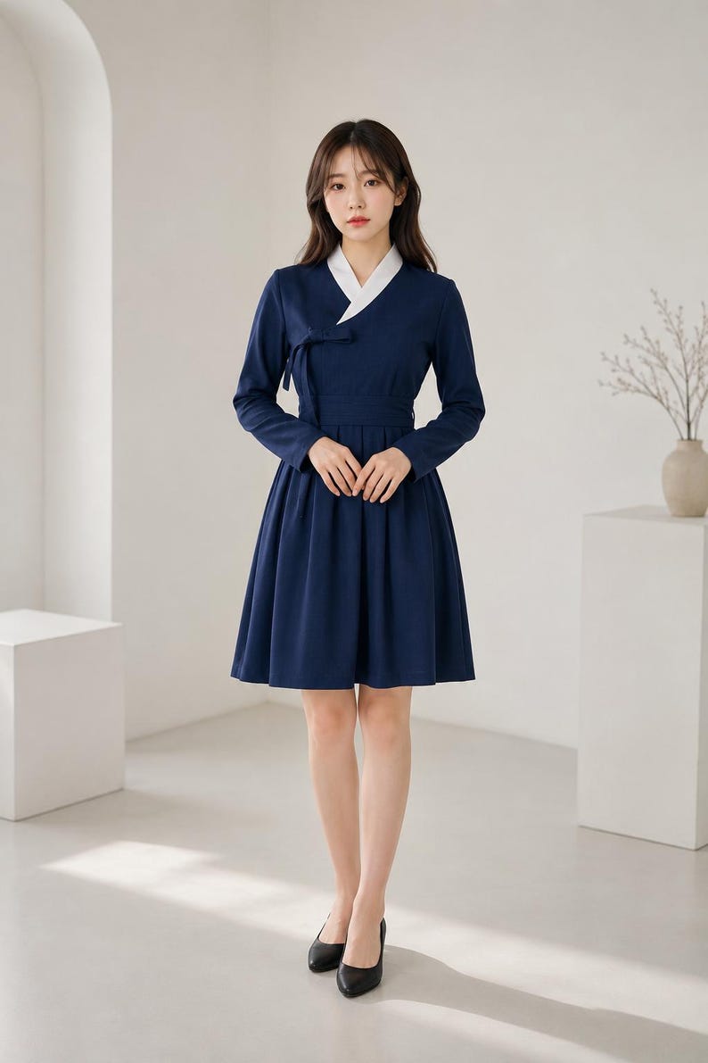 May include: A navy blue knee-length dress featuring a white V-neck collar, long sleeves, and a wrap-style bodice with a side tie closure. The skirt is pleated and flares from the waist, paired with black closed-toe pumps for a formal appearance.