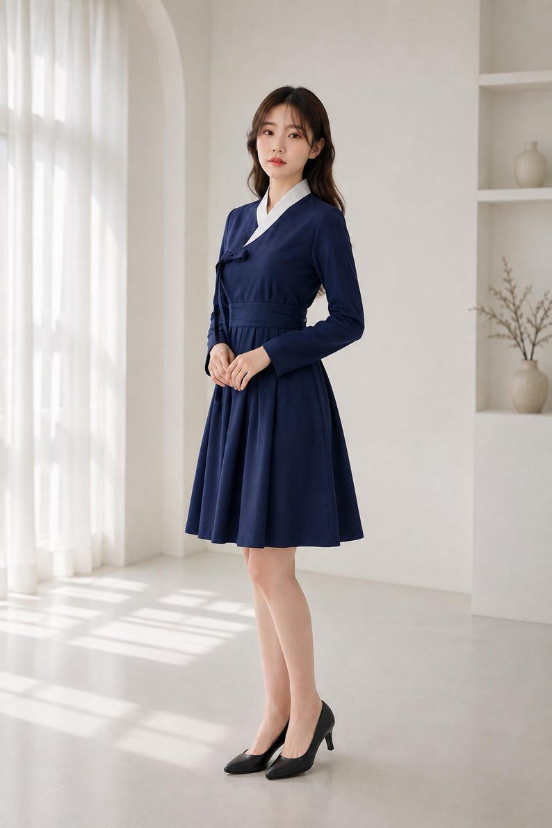 May include: A navy blue knee-length dress with long sleeves, a white V-neck collar, and a wrap-style bodice featuring a side tie closure. The garment has a fitted waist and a pleated skirt that falls to the knees, paired with black high heels.