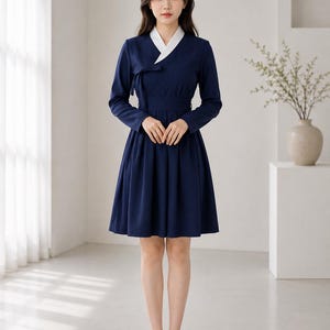 May include: A navy blue knee-length dress featuring a white V-neck collar, long sleeves, and a wrap-style bodice with a side tie closure. The skirt is pleated, creating a flared silhouette, and the garment is paired with black pointed-toe heels.