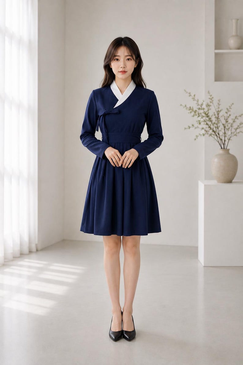 May include: A navy blue knee-length dress featuring a white V-neck collar, long sleeves, and a wrap-style bodice with a side tie closure. The skirt is pleated, creating a flared silhouette, and the garment is paired with black pointed-toe heels.