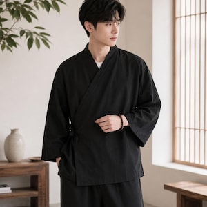 May include: A black wrap-style shirt with long sleeves and a V-neckline, featuring a side tie closure. The garment is constructed from a solid black fabric with a subtle texture, paired with matching black trousers in a relaxed, straight-leg cut.