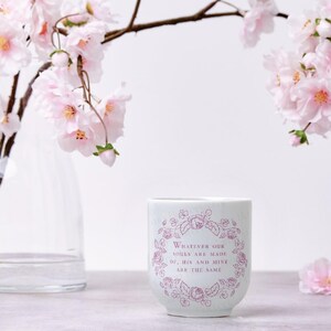 Puede incluir: Un vaso de cer&aacute;mica blanco con un dise&ntilde;o floral y la cita, "Whatever our souls are made of, his and mine are the same." Flores de cerezo rosas y un jarr&oacute;n transparente en el fondo.
